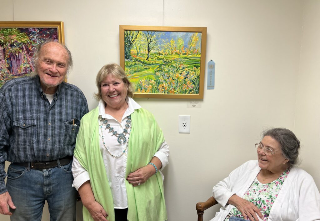 An artist smiling beside her award-ribboned landscape painting with two guests at an exhibition