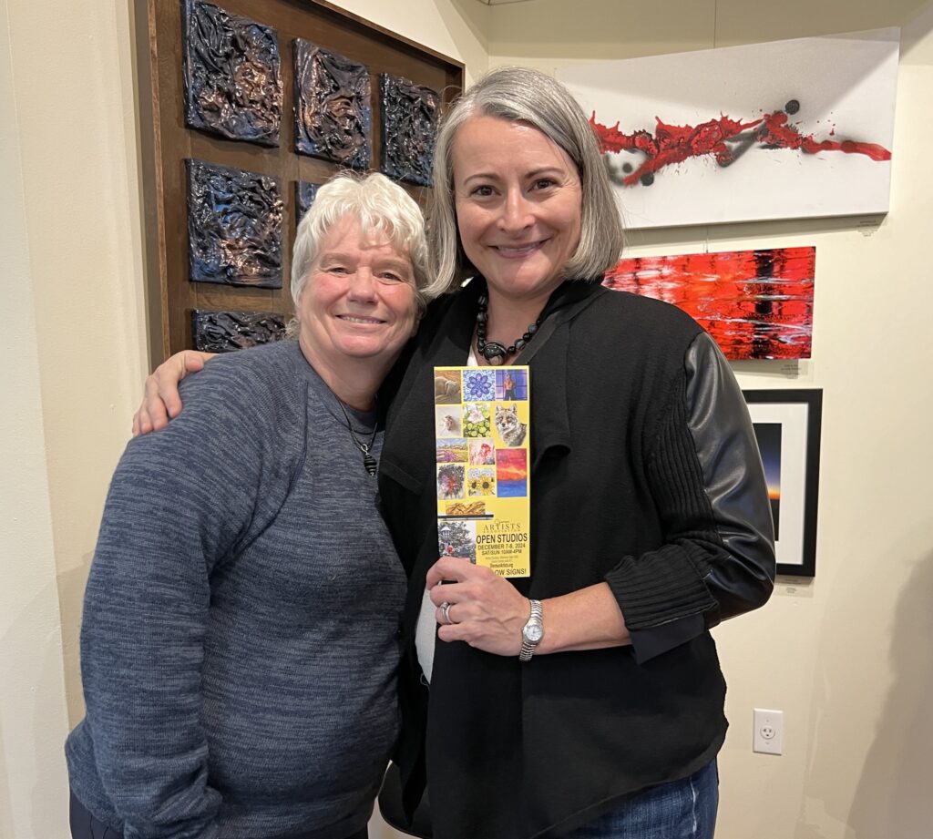 Two smiling friends holding an Open Studios brochure in front of artwork