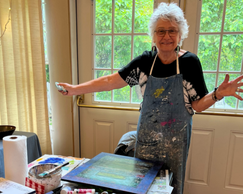 Artist Carolyn Cohen smiling in her paint-spattered apron beside a painting in her home studio